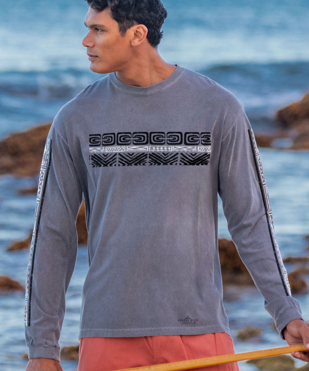 A man wearing a long sleeve crewneck t-shirt with tribal patterns printed across the chest, standing by the sea. View 2