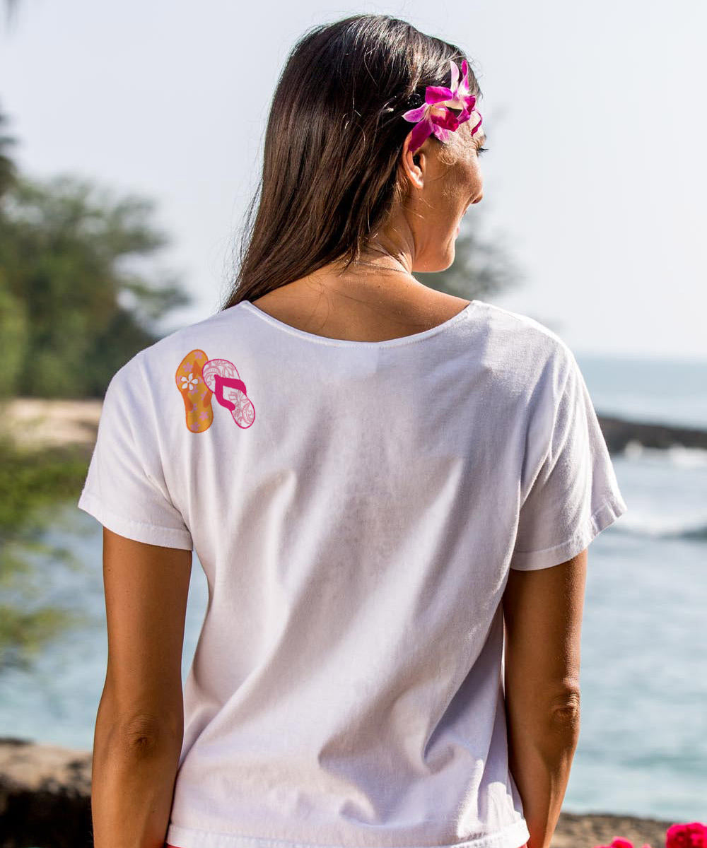 woman standing on the beach wearing a white t-shirt with flip-flops on it. View 3
