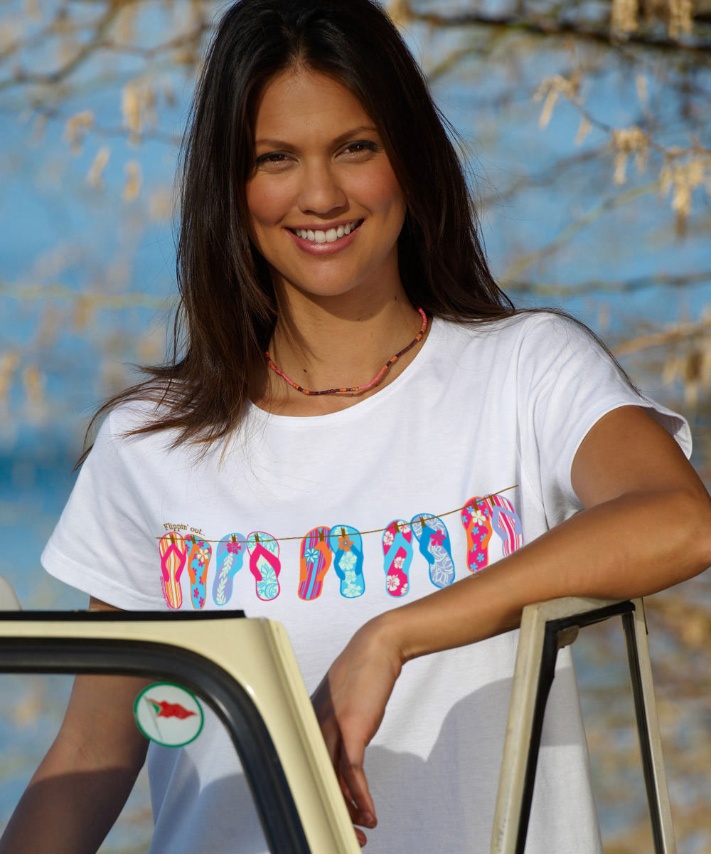 woman standing on the beach wearing a white t-shirt with flip-flops on it. View 2