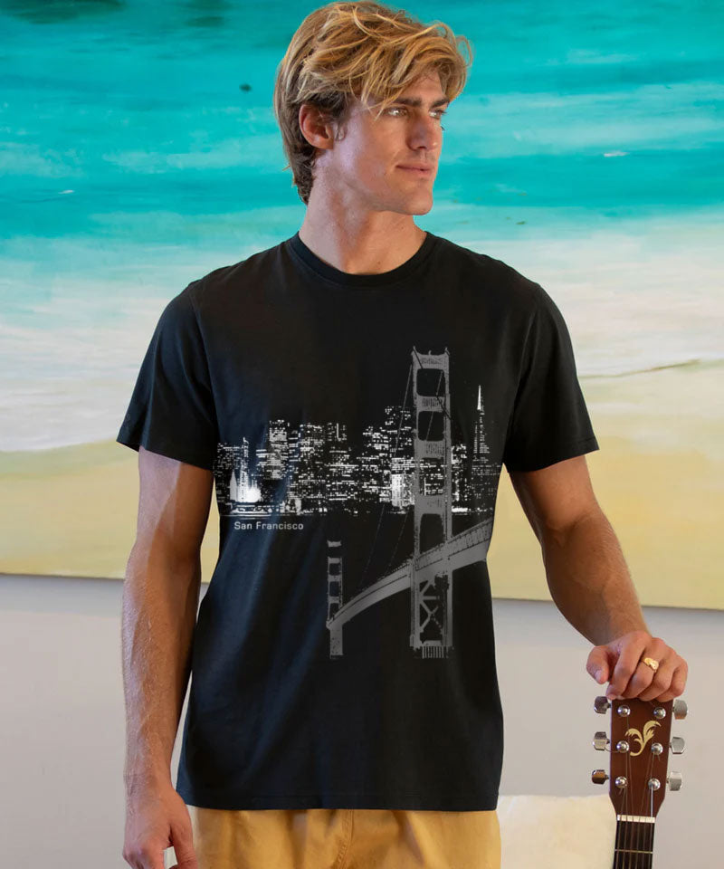 Man wearing a black t-shirt with a cityscape design, holding a guitar, against a beach background. View 2