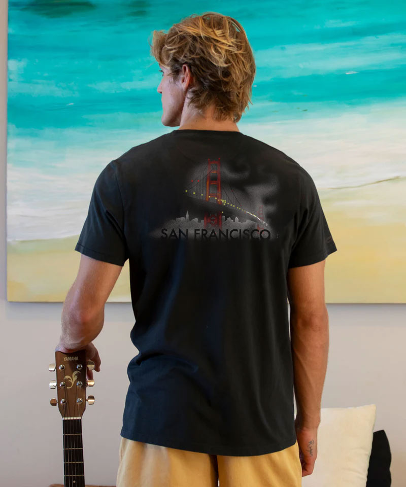 Person wearing a black t-shirt with 'San Francisco' design, standing in front of a scenic painting. View 2