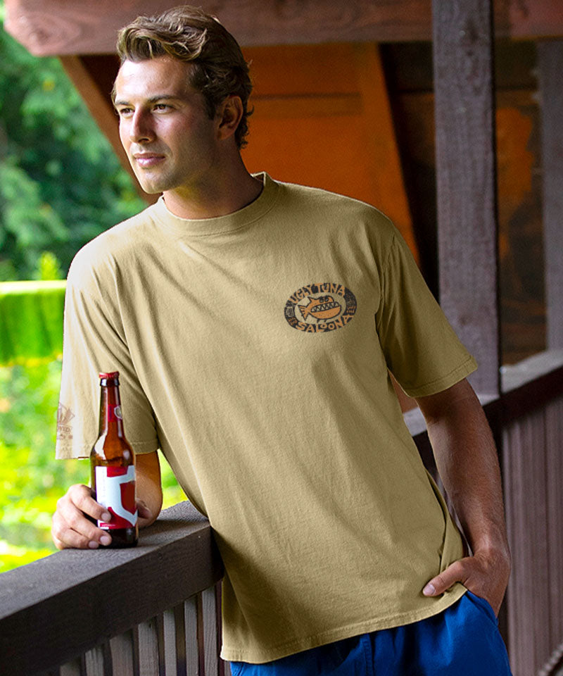 Man wearing a beige t-shirt with a logo, holding a beer bottle on a balcony. View 3
