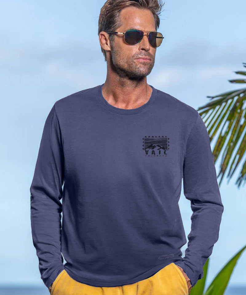 Man wearing a navy long-sleeve shirt with a logo, sunglasses, and yellow shorts against a beach background. View 3