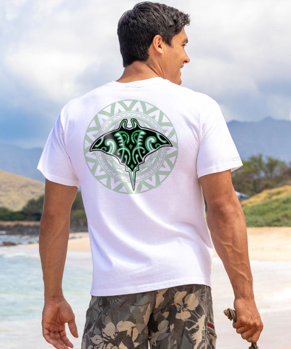 A man facing away from the camera wears a white short sleeve crewneck t-shirt with a tribal manta ray design on the back, standing on a beach. View 2