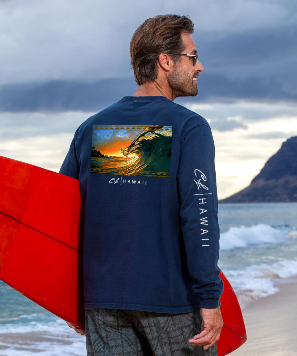 A man standing on a beach holding a red surfboard, wearing a navy long sleeve crewneck t-shirt with a graphic of a sunset wave and the text 'Hawaii'. View 2