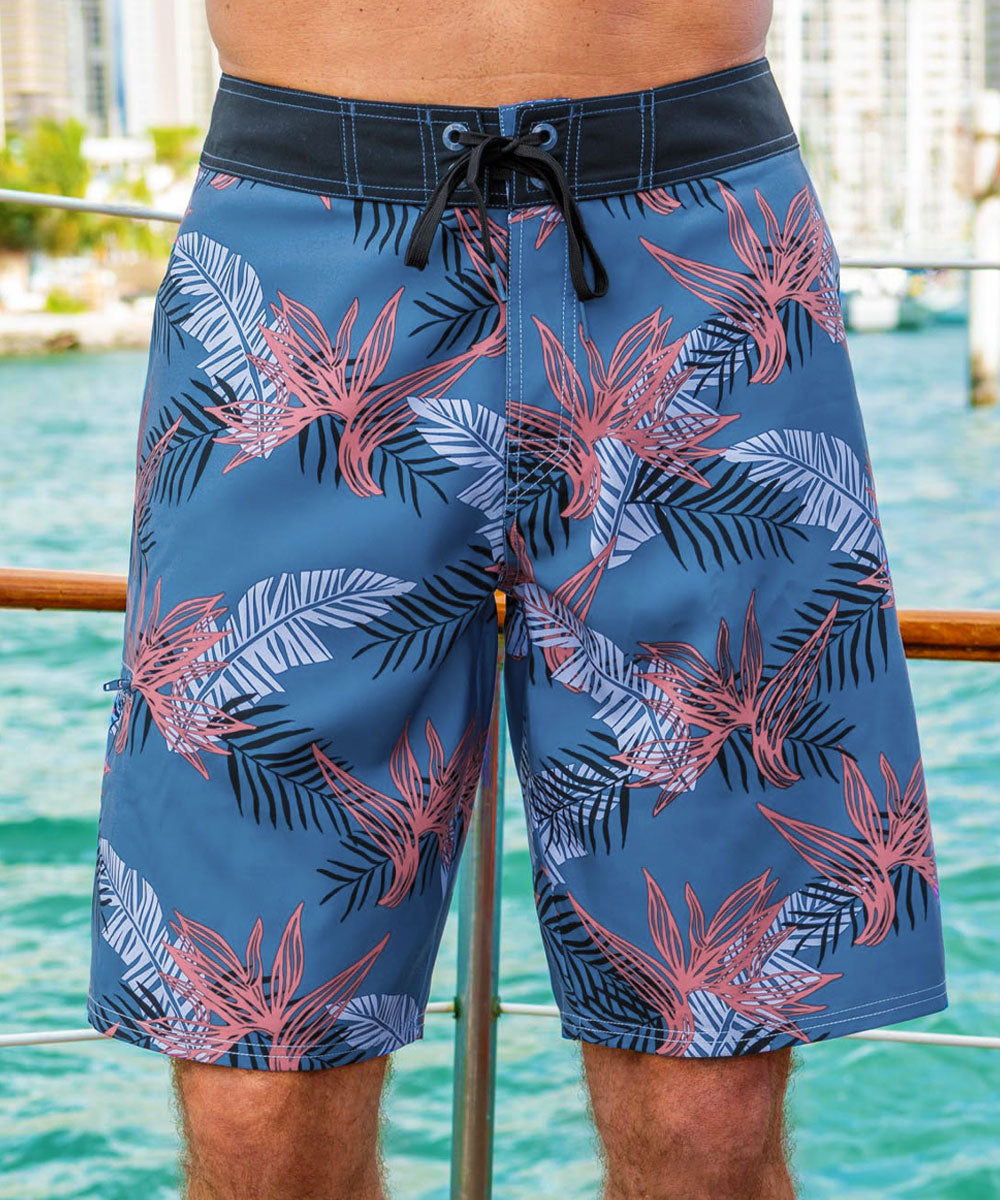 A person wearing a pair of slate blue board shorts with a tribal pattern, displayed in a beach setting. View 2