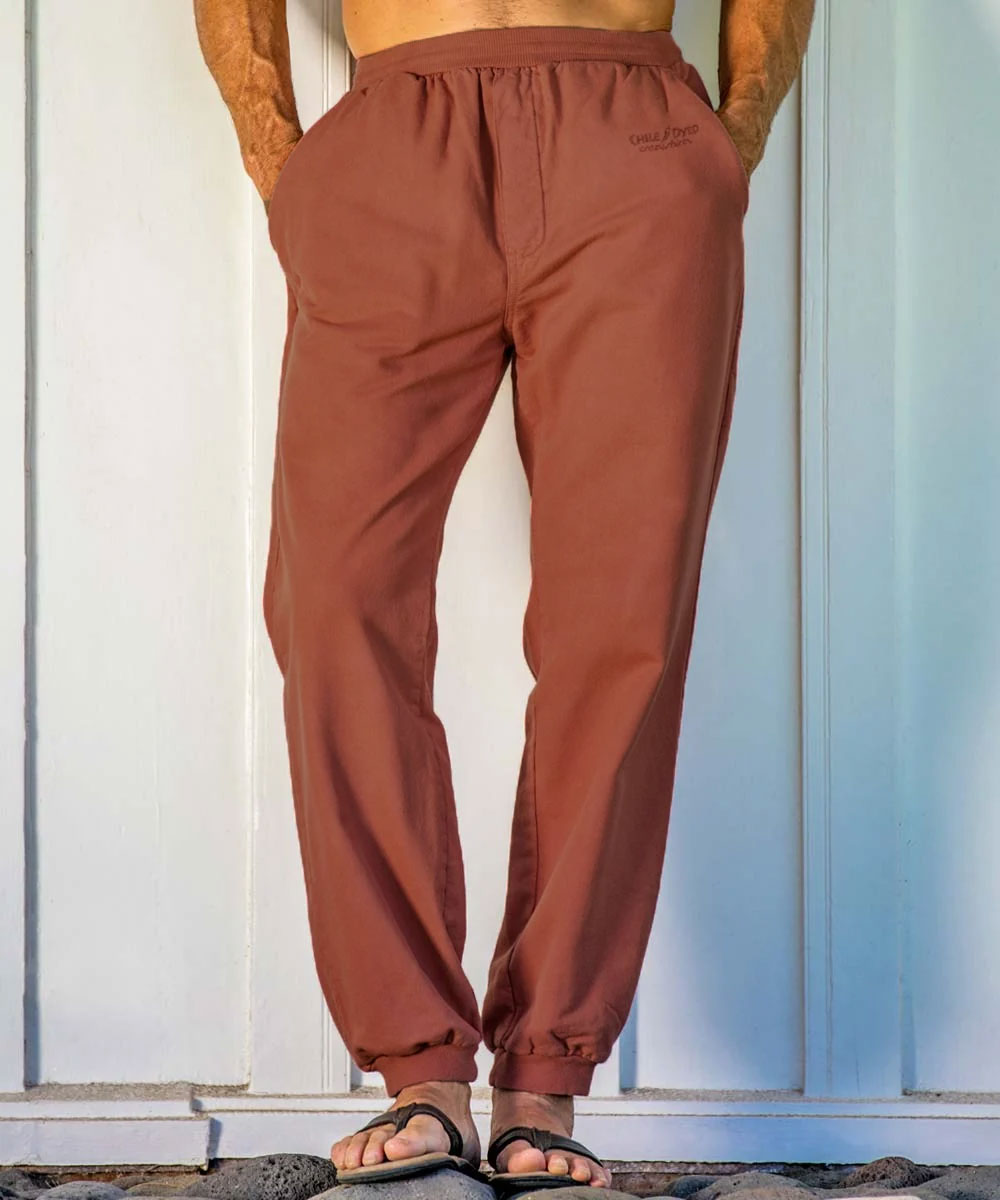 A person standing by a light blue wall wearing rust pants and black slippers, with hands in pockets. View 2