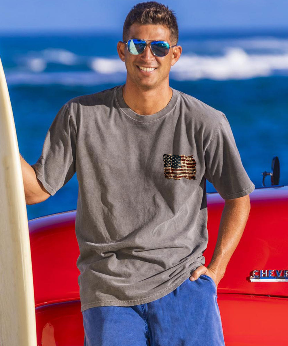 A man facing the camera wearing a grey t-shirt with a small American flag graphic printed on the chest, holding a surfboard and the beach in the background. View 3