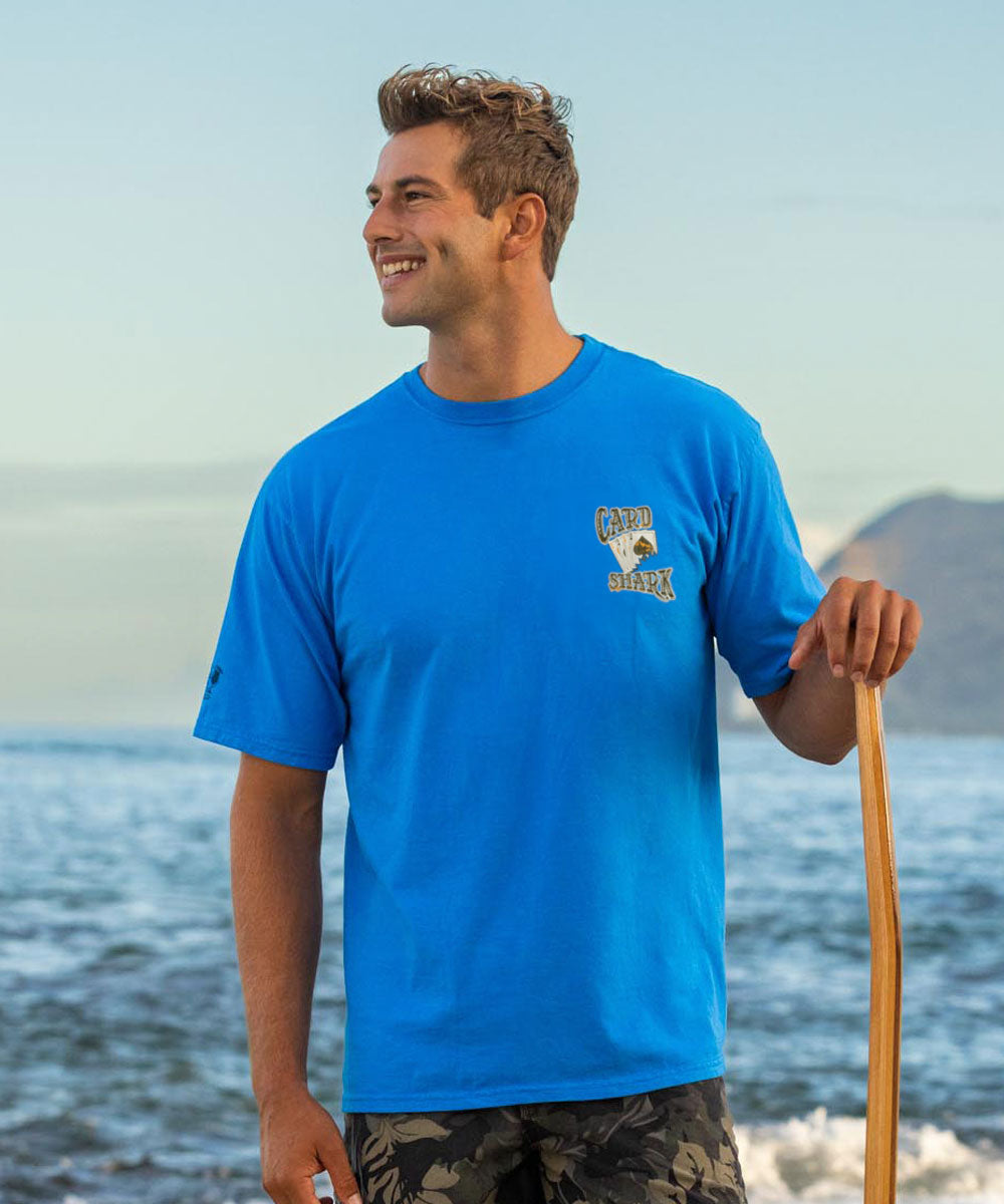 Man wearing a blue t-shirt with a logo, holding a paddle, standing by the ocean. View 3