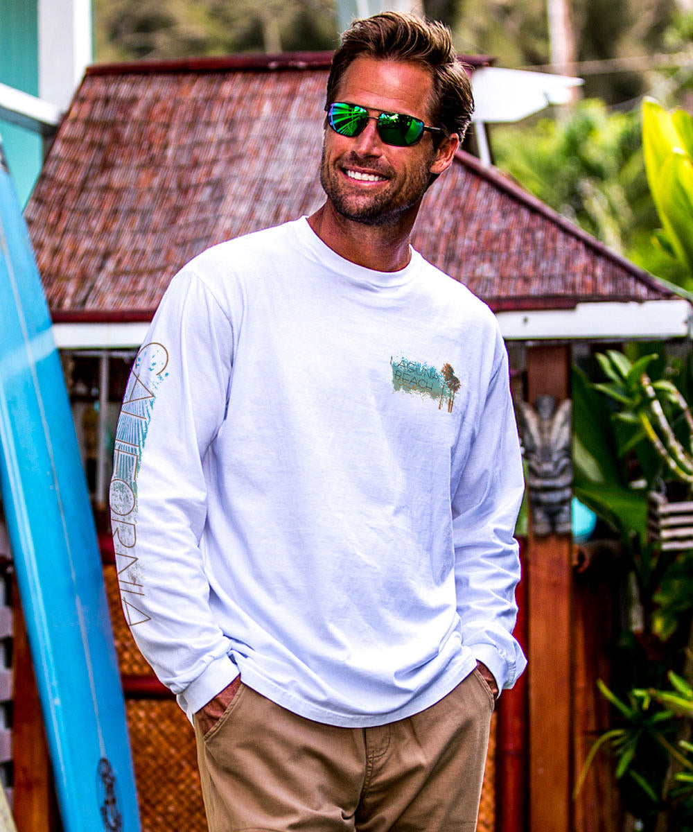 Man wearing a white long-sleeve shirt with a logo, standing in front of a wooden building and surfboard. View 3