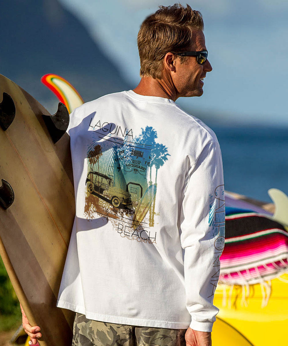 Man wearing a white long-sleeve shirt with a graphic design, holding surfboards on a beach. View 2