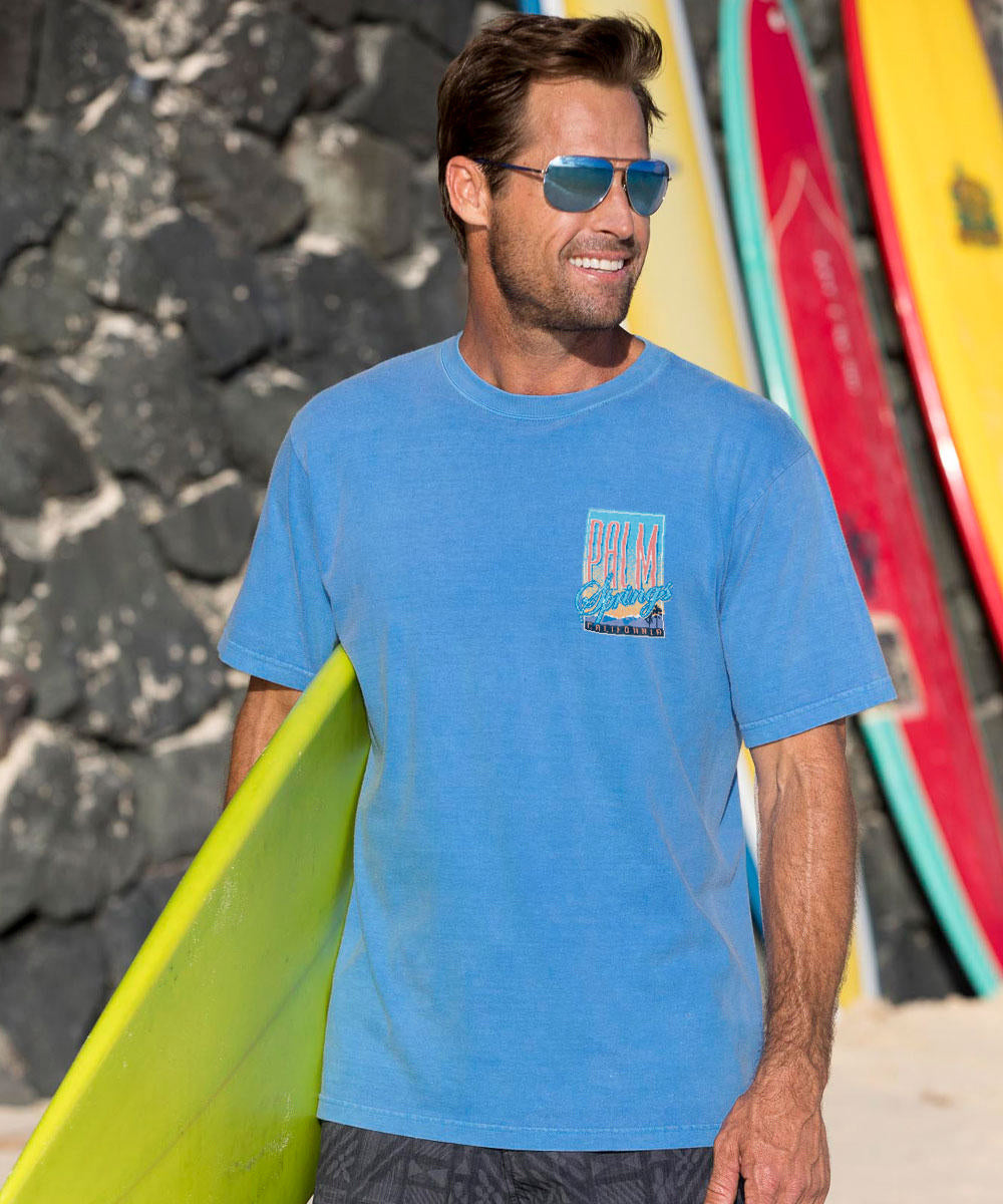 Man in blue t-shirt holding a yellow surfboard with colorful boards in the background. View 3