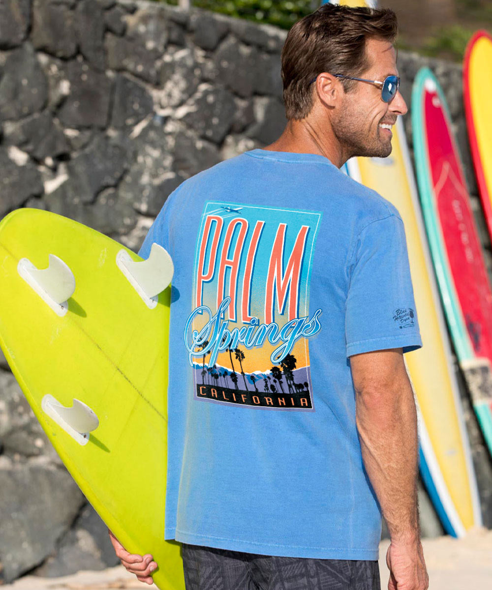 Man in blue t-shirt with palm tree graphic holding a yellow surfboard. View 2