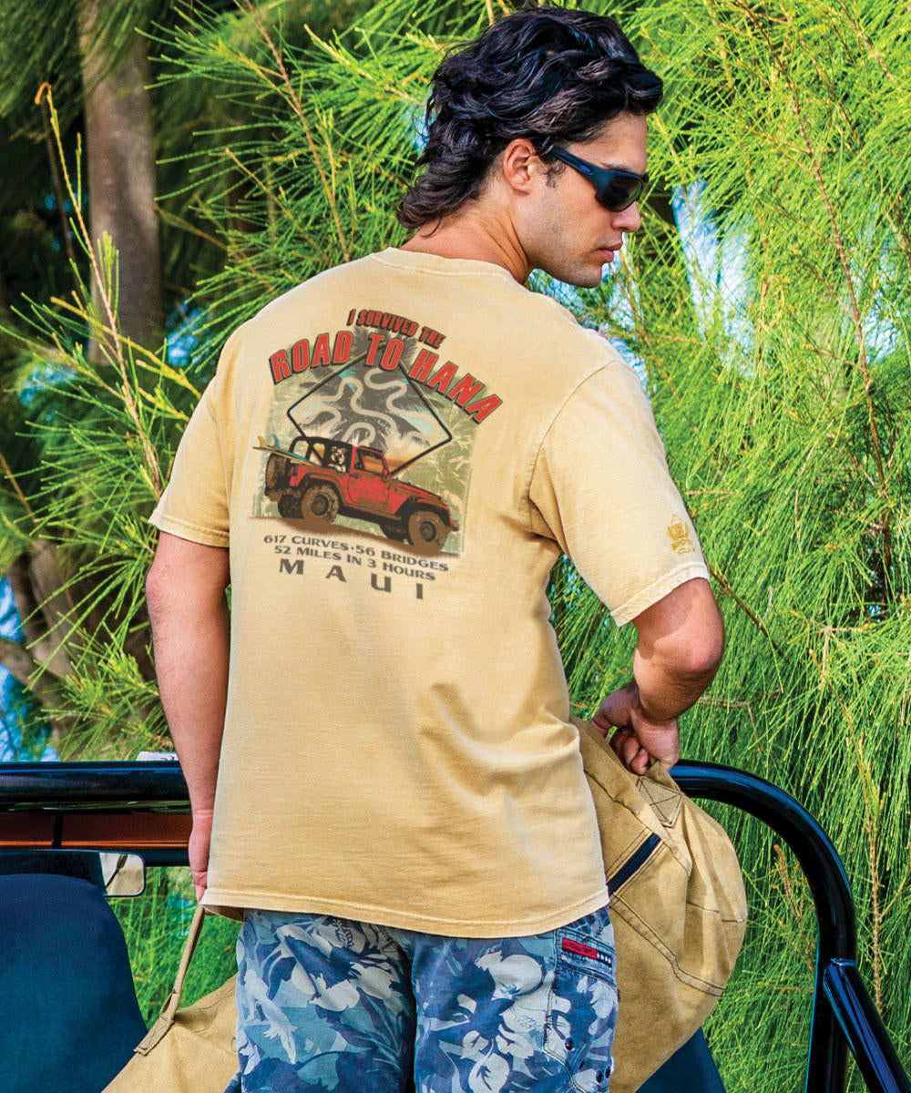 Man wearing a beige t-shirt with a graphic design, standing outdoors with greenery in the background. View 2