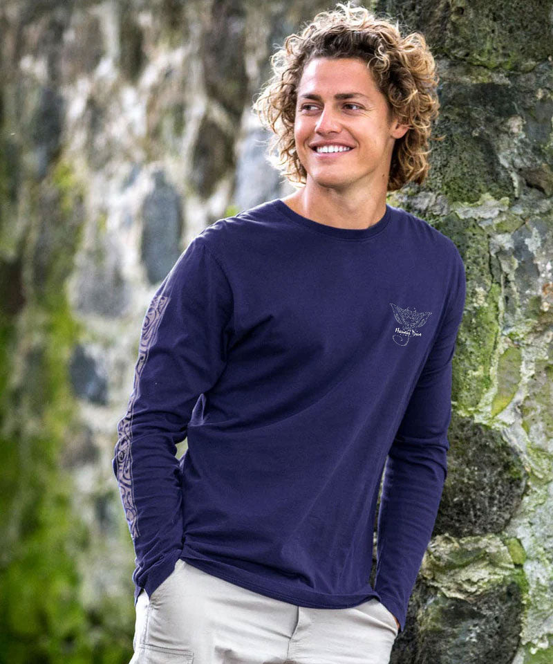 Man wearing a navy blue long-sleeve shirt with a logo, standing against a stone wall. View 3