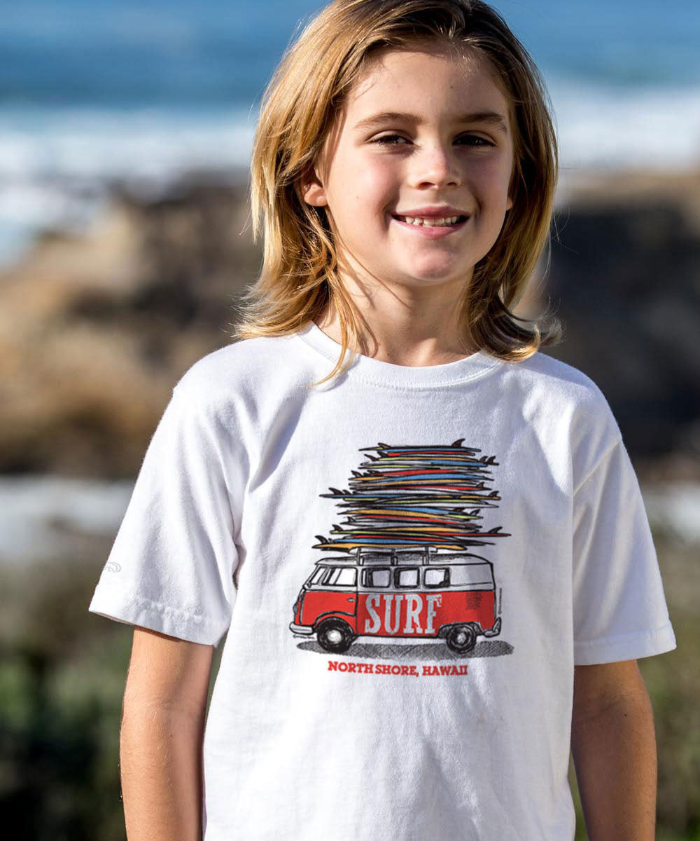 A smiling young boy with blonde hair stands outdoors on a beach wearing a white graphic t-shirt. The t-shirt features a red and white surf van carrying a tall stack of surfboards, with 'SURF' written on the side and 'NORTH SHORE, HAWAII' printed below. View 2