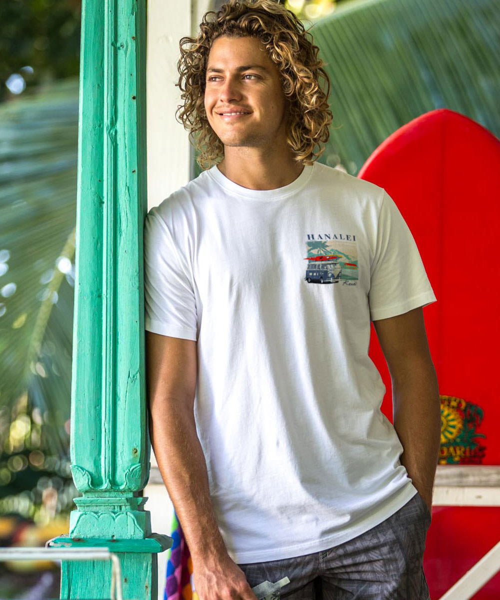 Man wearing a white t-shirt with a graphic design, standing next to a red surfboard indoors. View 3