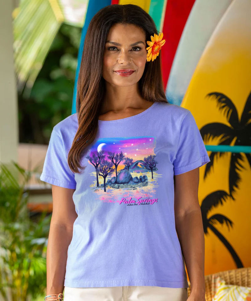 Woman wearing a purple t-shirt with a graphic design in front of colorful surfboards. View 2