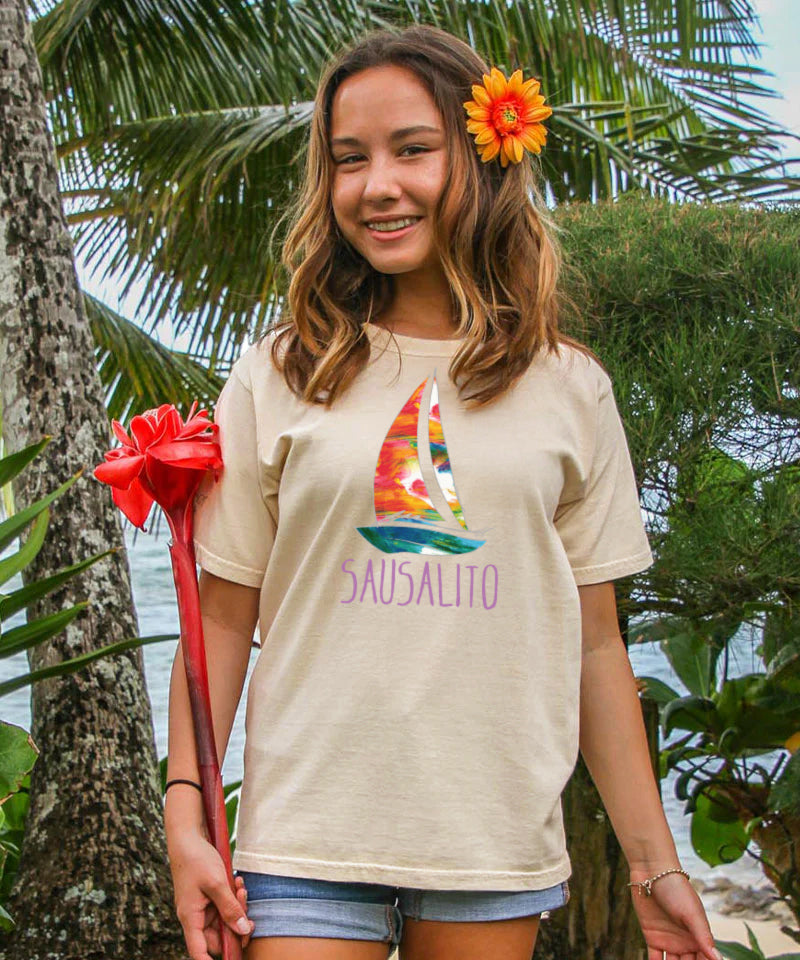 Young girl wearing a t-shirt with a colorful sailboat design and 'Sausalito' text, standing in a tropical setting. View 2