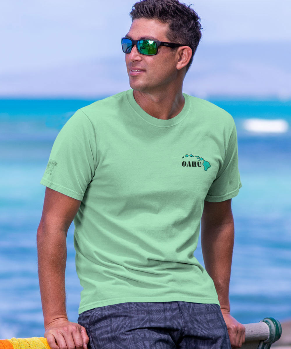Man wearing a light green t-shirt with a logo, standing by the ocean. View 3