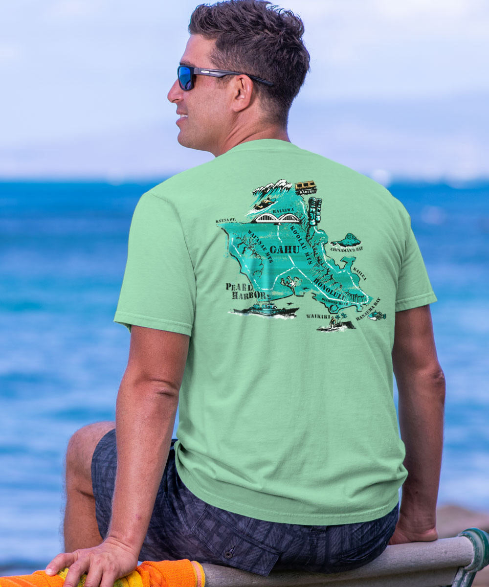 Man wearing a green t-shirt with a graphic design, sitting by the ocean. View 2