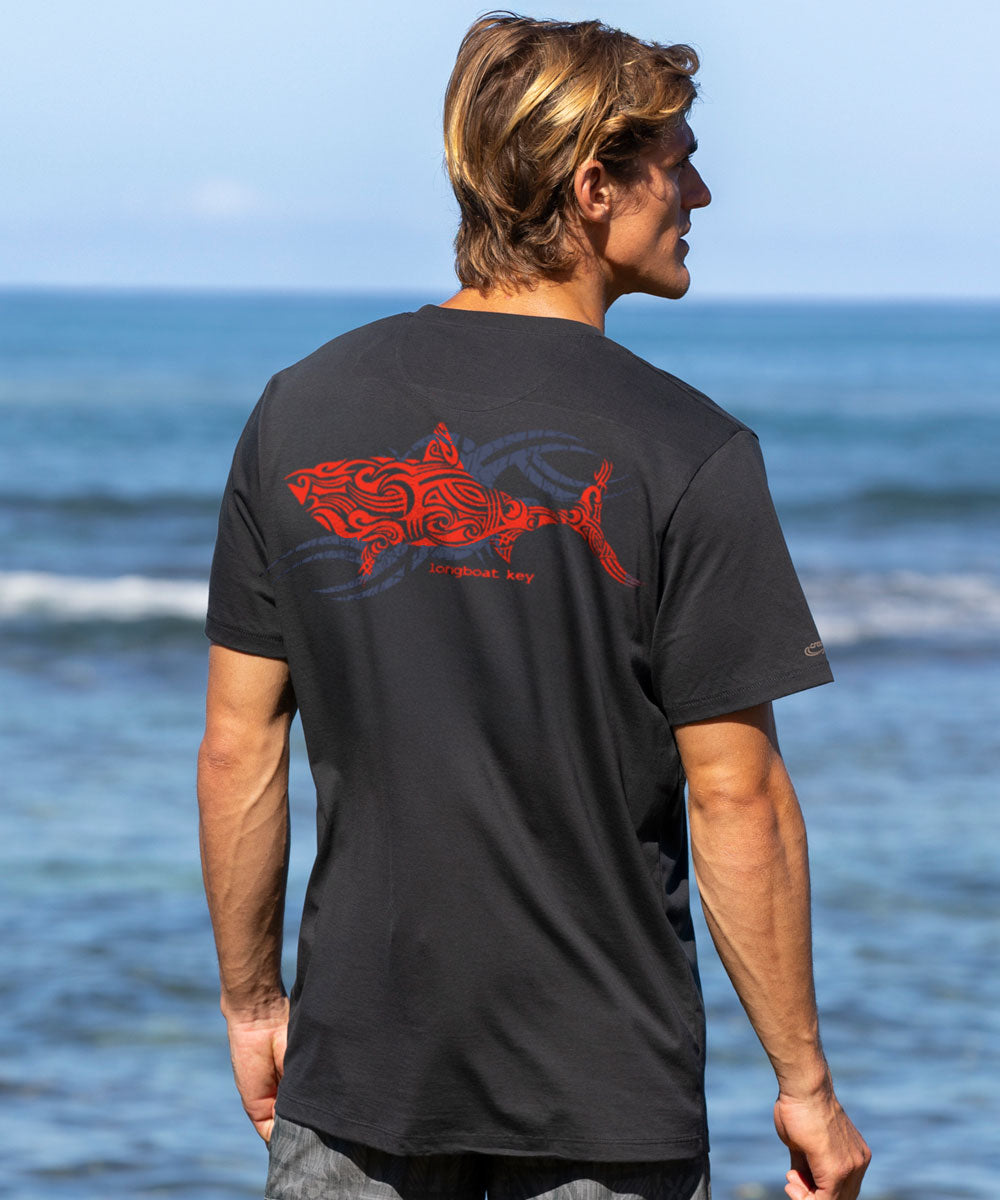 Man wearing a black t-shirt with a red shark design on the back, standing by the ocean. View 2
