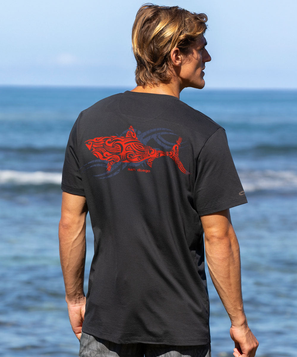 Man wearing a black t-shirt with a red shark design on the back, standing by the ocean. View 2