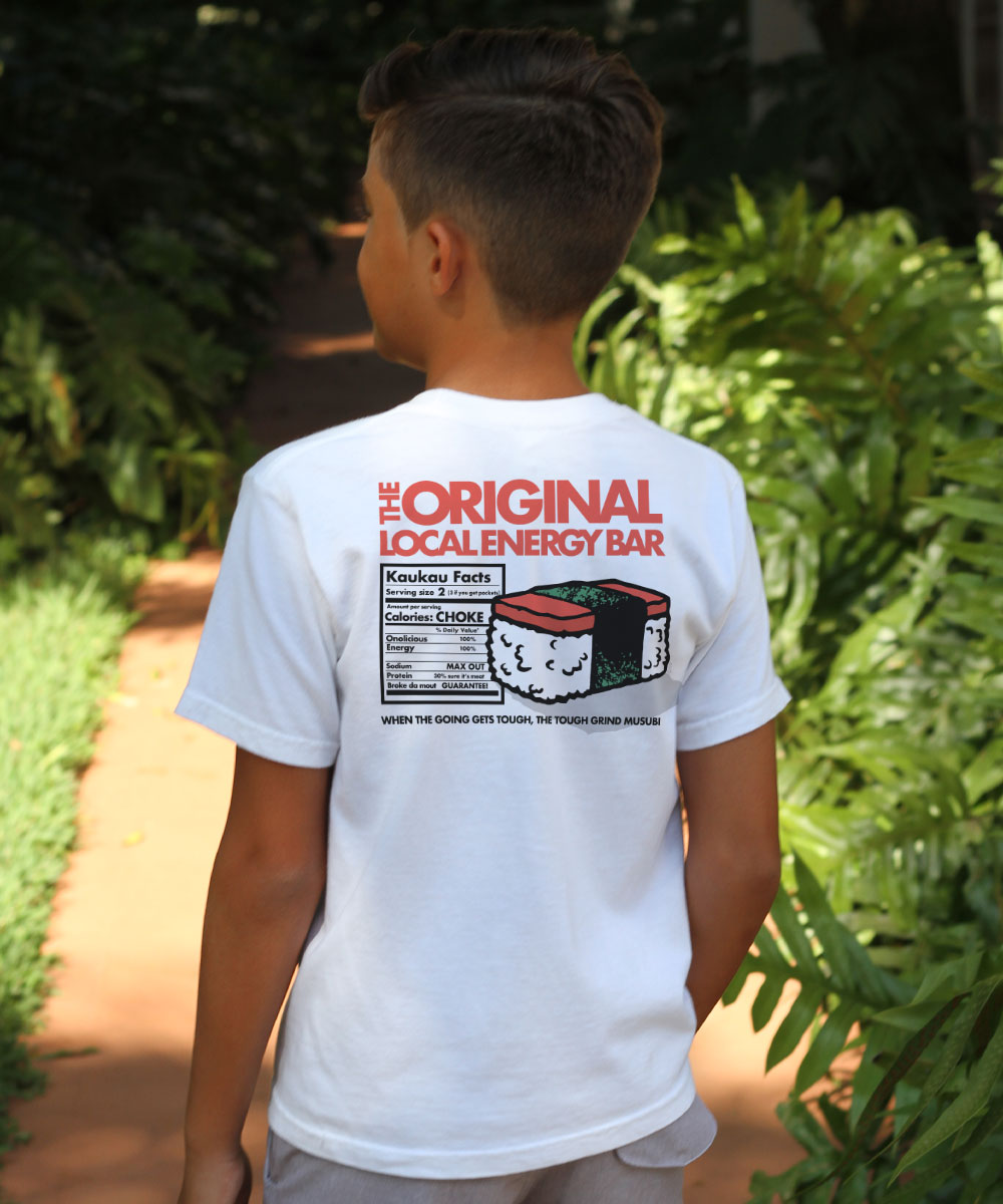 A child facing away from camera, wearing a white t-shirt with a graphic design of a spam musubi and text of the nutritional facts in a joking manner, greenery in background. View 2