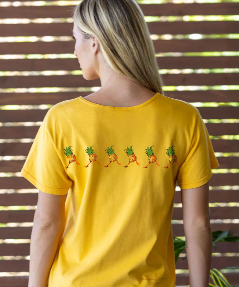 Woman facing away from the camera, wearing a yellow t-shirt with pineapple and rooster design across the back, against a wooden slat background View 3