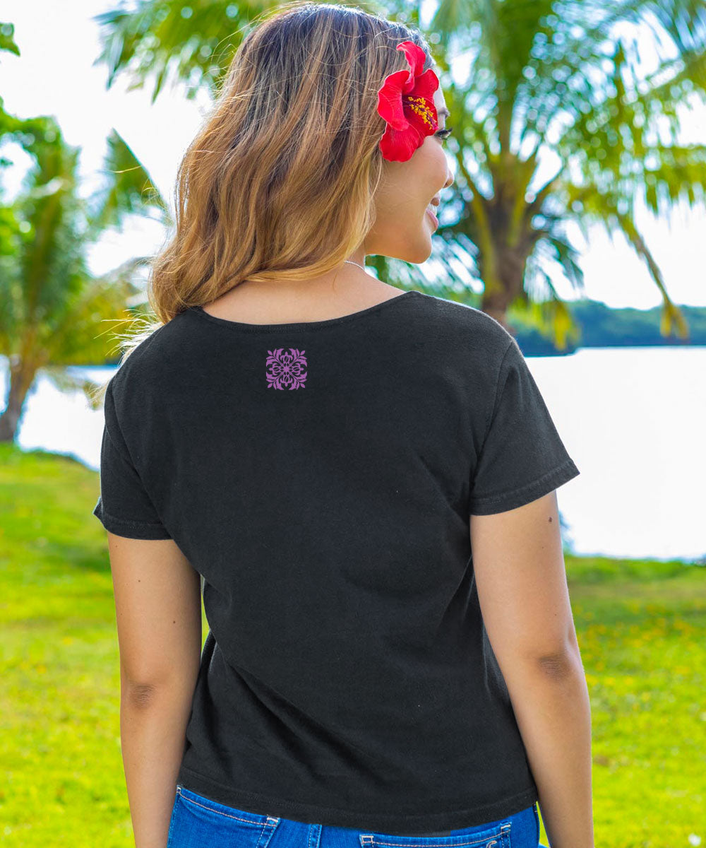 A woman facing away from the camera, wearing a black scoop neck t-shirt with a small purple Quilt floral design on the top middle area with palm trees and water in background. View 3