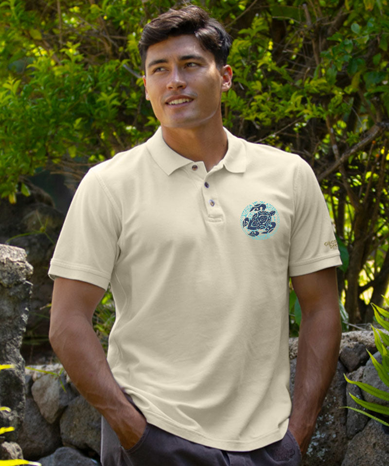 A smiling man wearing a beige short sleeve polo shirt with a turtle design on the chest and logo faux horn buttons, standing in front of a tropical setting. View 2