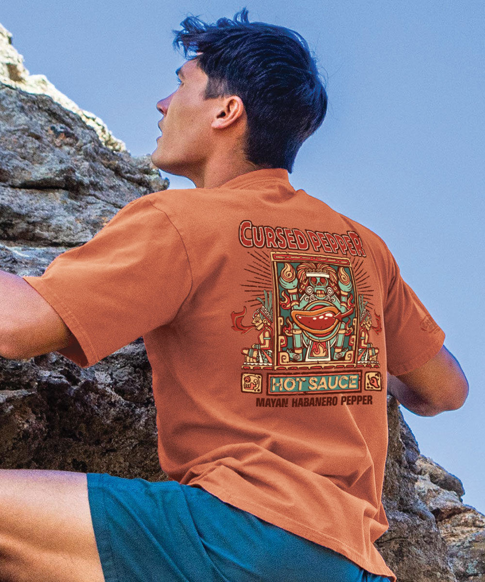 A person wearing a brown short sleeve crewneck t-shirt with 'Cursed Pepper' text and graphics on the back. View 2
