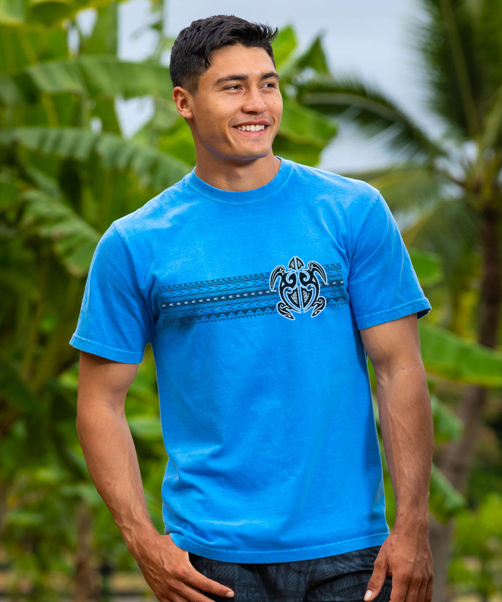 A person wearing a blue short-sleeve crewneck t-shirt with a tribal turtle design, standing in front of a tropical backdrop. View 2