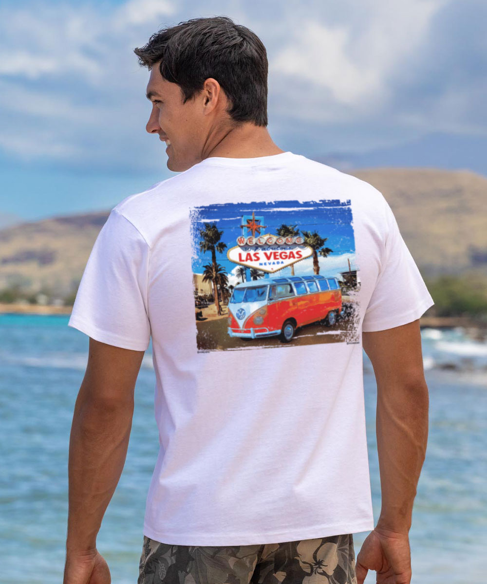 Man wearing a white t-shirt with a graphic of a van and Las Vegas sign, standing by a beach. View 2