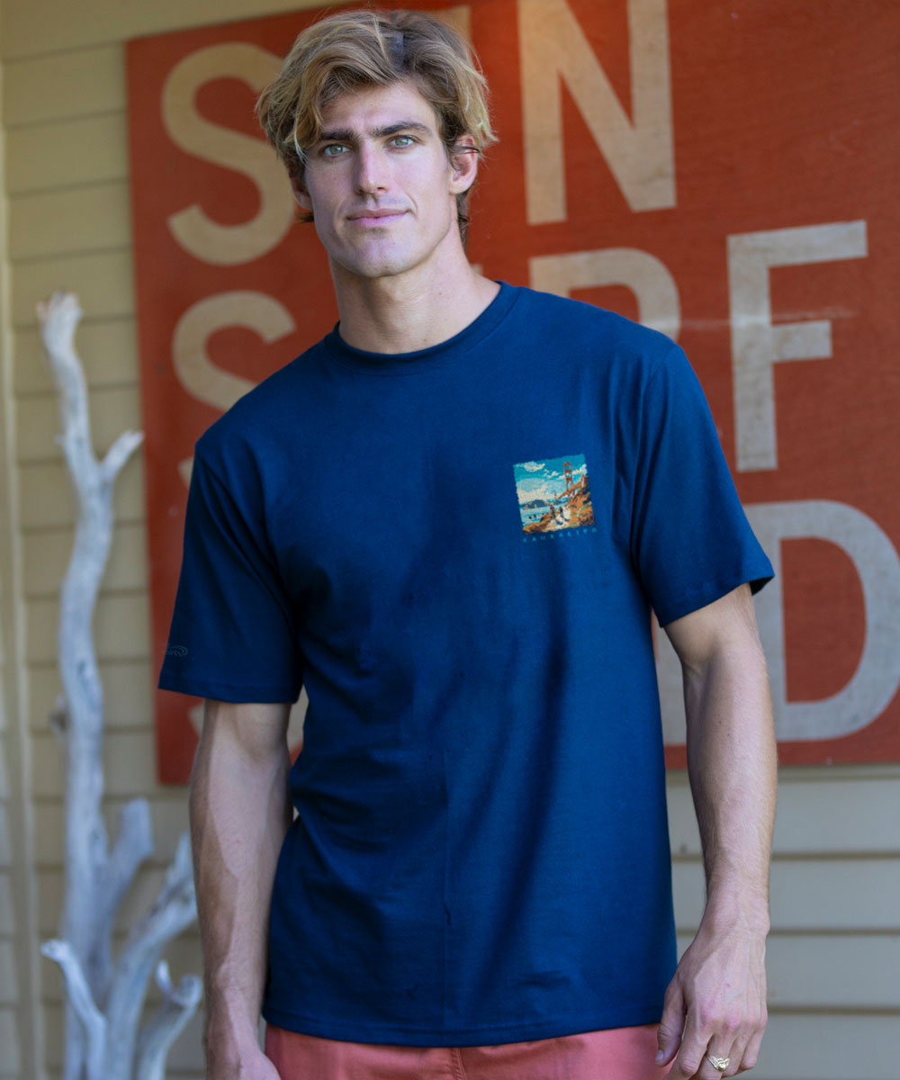 Man wearing a blue t-shirt with a small graphic on the chest, standing in front of a red sign. View 3