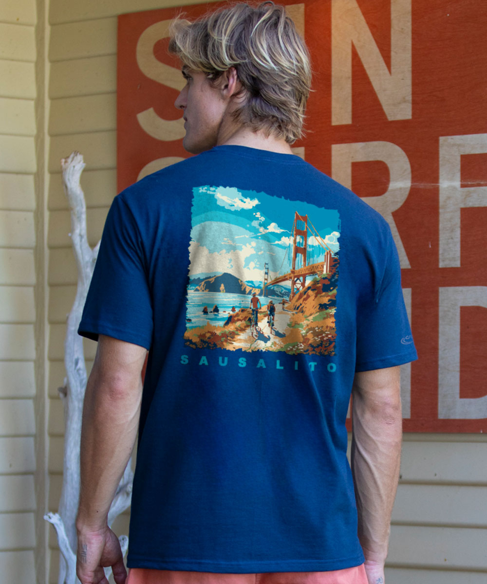 Person wearing a blue t-shirt with a scenic print, standing in front of a building with large letters. View 2