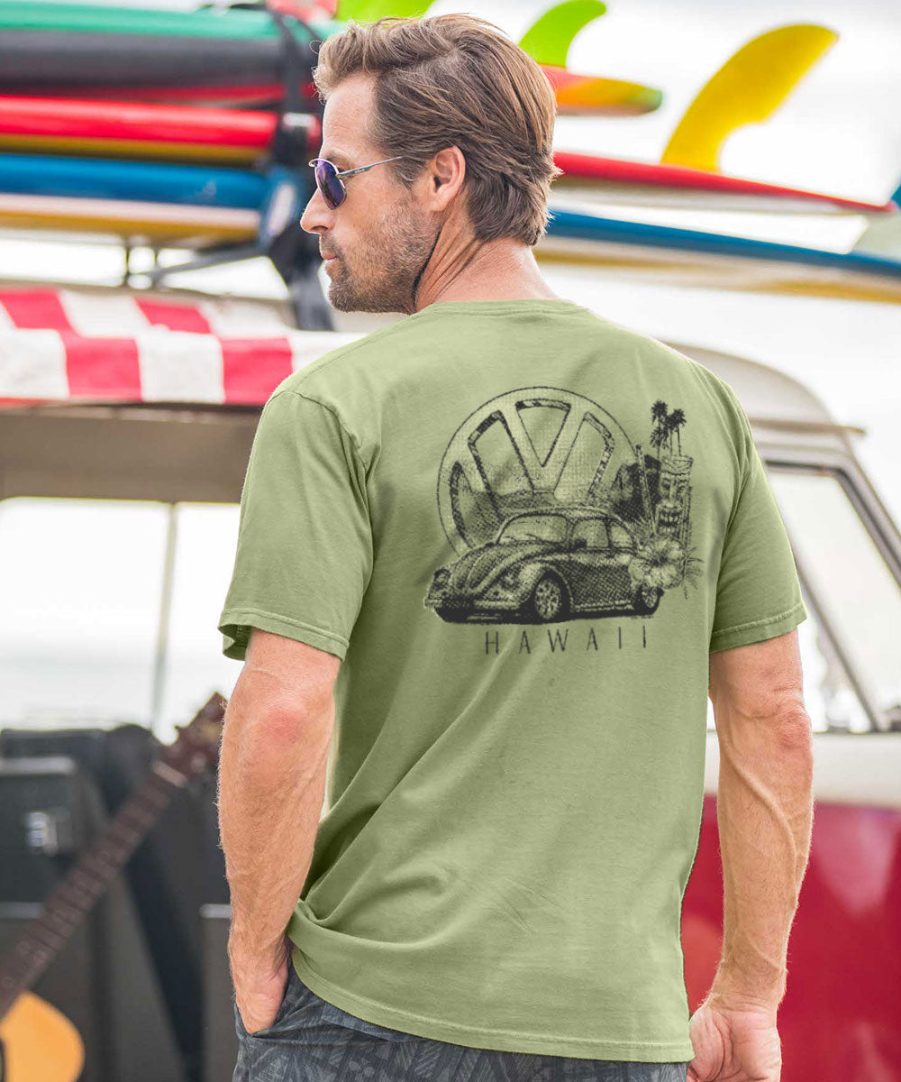 A man wearing a green short sleeve crewneck t-shirt with a Volkswagen and Hawaiian imagery on the back, standing near a colorful boat. View 2