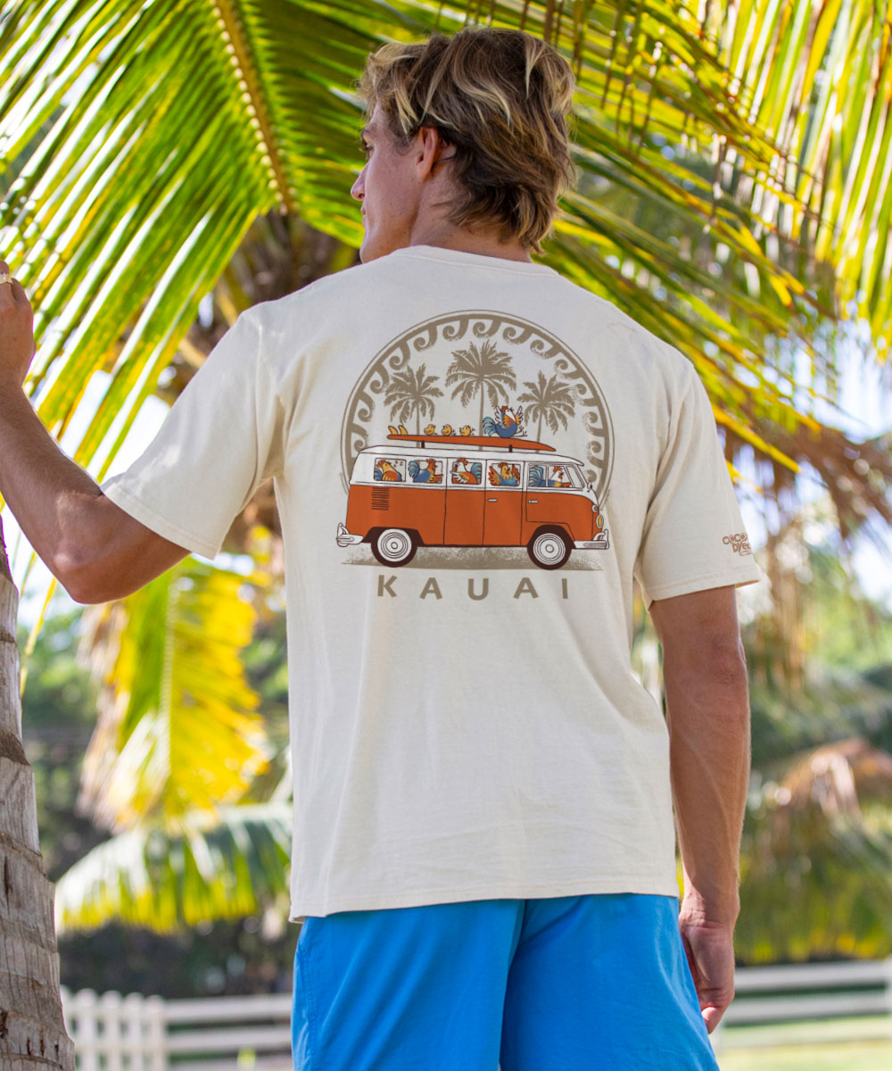 A person wearing a white short sleeve crewneck t-shirt with a graphic of a chicken van and the text 'Kauai' below it, standing under palm trees. View 2