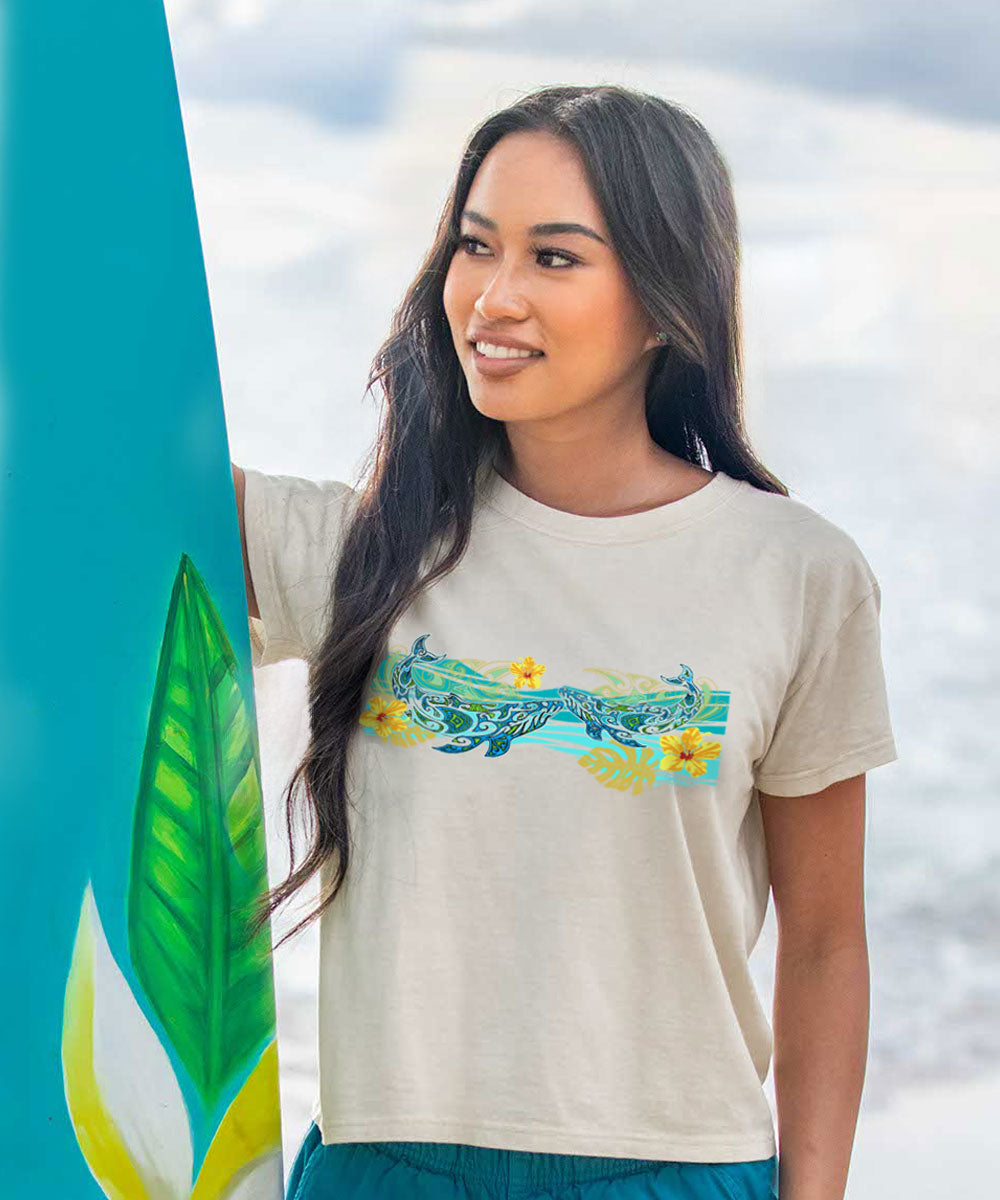 A woman wearing a beige short sleeve crewneck t-shirt with a tribal whale design in front, standing in front of a surfboard. View 2