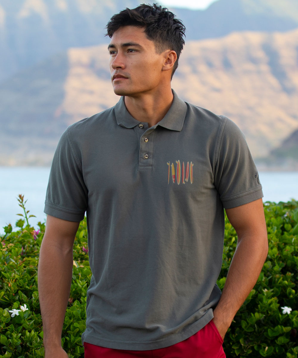 Man wearing a gray polo shirt with a logo, standing outdoors with mountains and water in the background. View 2