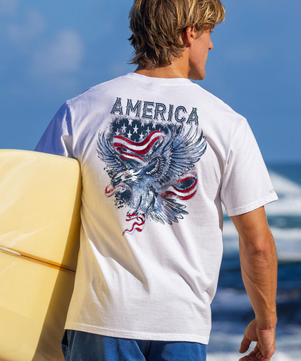 A person wearing a white short sleeve crewneck t-shirt with an eagle and the text 'AMERICA' on the back, standing in front of a beach scene. View 2