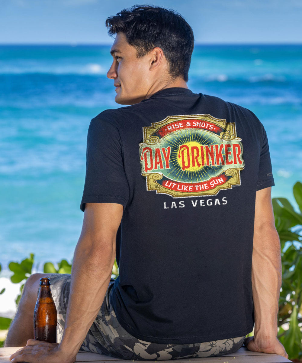 A person sitting with their back towards the camera wearing a black short sleeve crewneck t-shirt with 'Day Drinker' text and graphics in a bold font along with '拉斯维加斯' (Las Vegas) written below. There is a bottle and a glass in front of the person on a surface, and the background features a beautiful beach and ocean. View 2