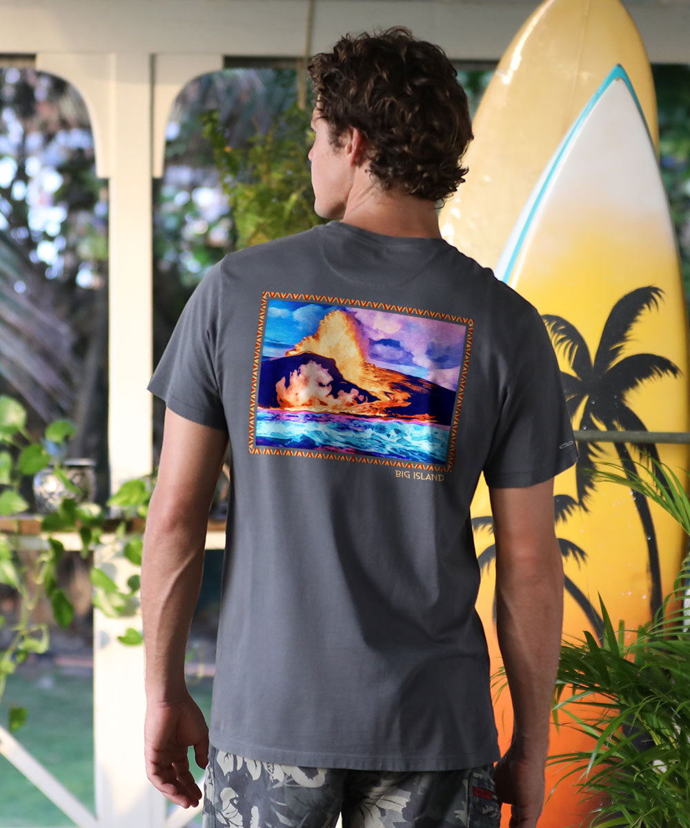 A person facing away from the camera wears a gray short-sleeve Pima t-shirt with a graphic of a volcanic eruption meeting the ocean, standing in front of a surfboard and tropical plants. View 2