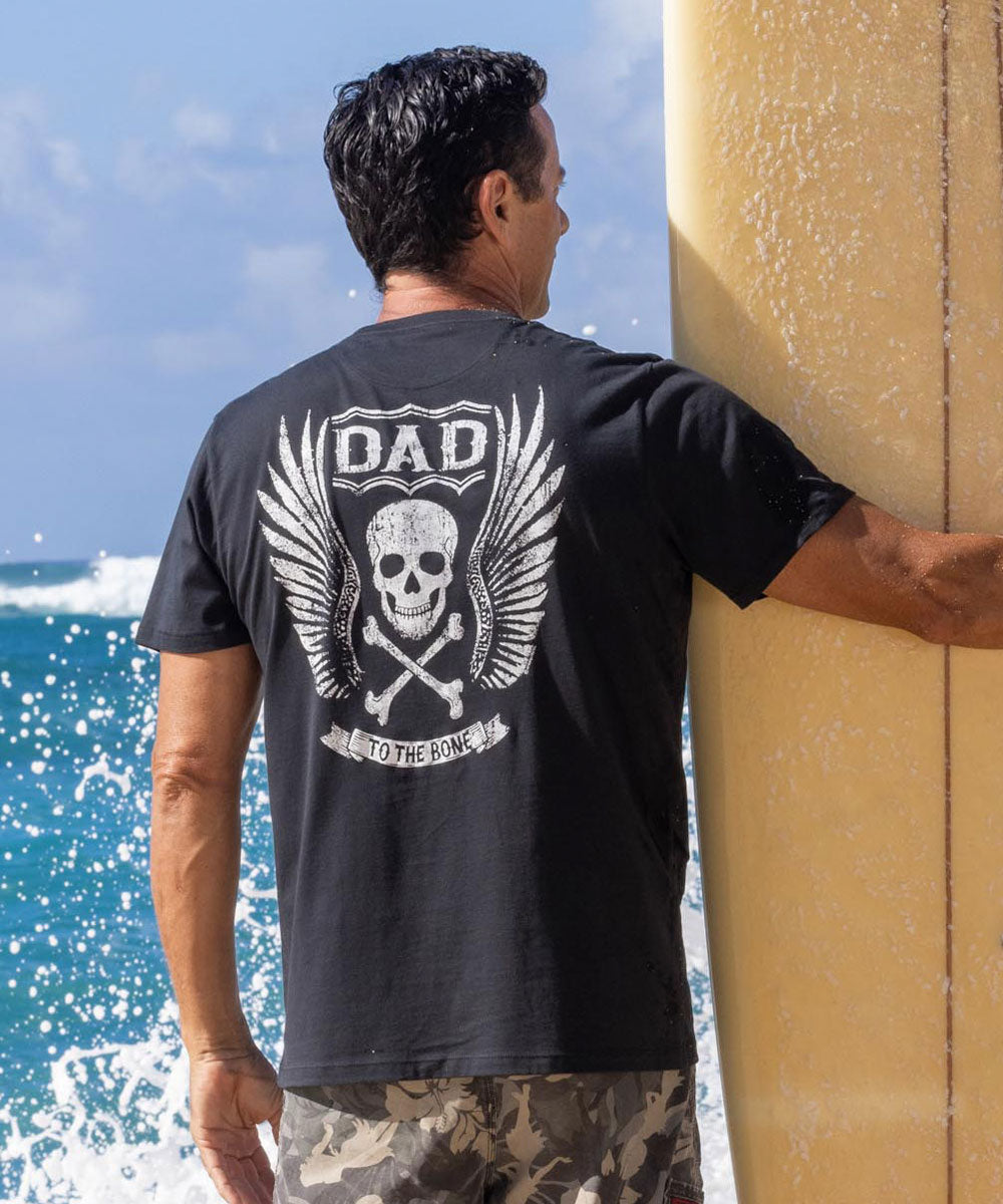 A person wearing a black short sleeve Pima cotton t-shirt with a graphic design on the back standing by a pole with the ocean in the background. View 2