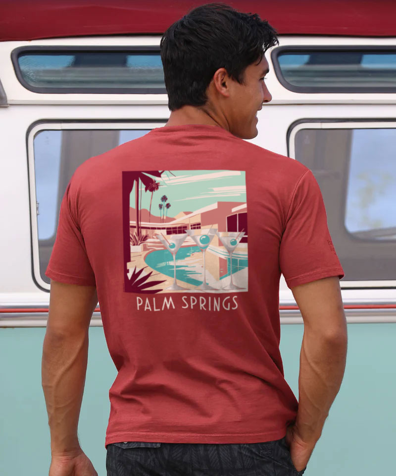 A person wearing a red short sleeve crewneck t-shirt with a graphic design of a building and text 'PALM SPRINGS' on the back. View 2