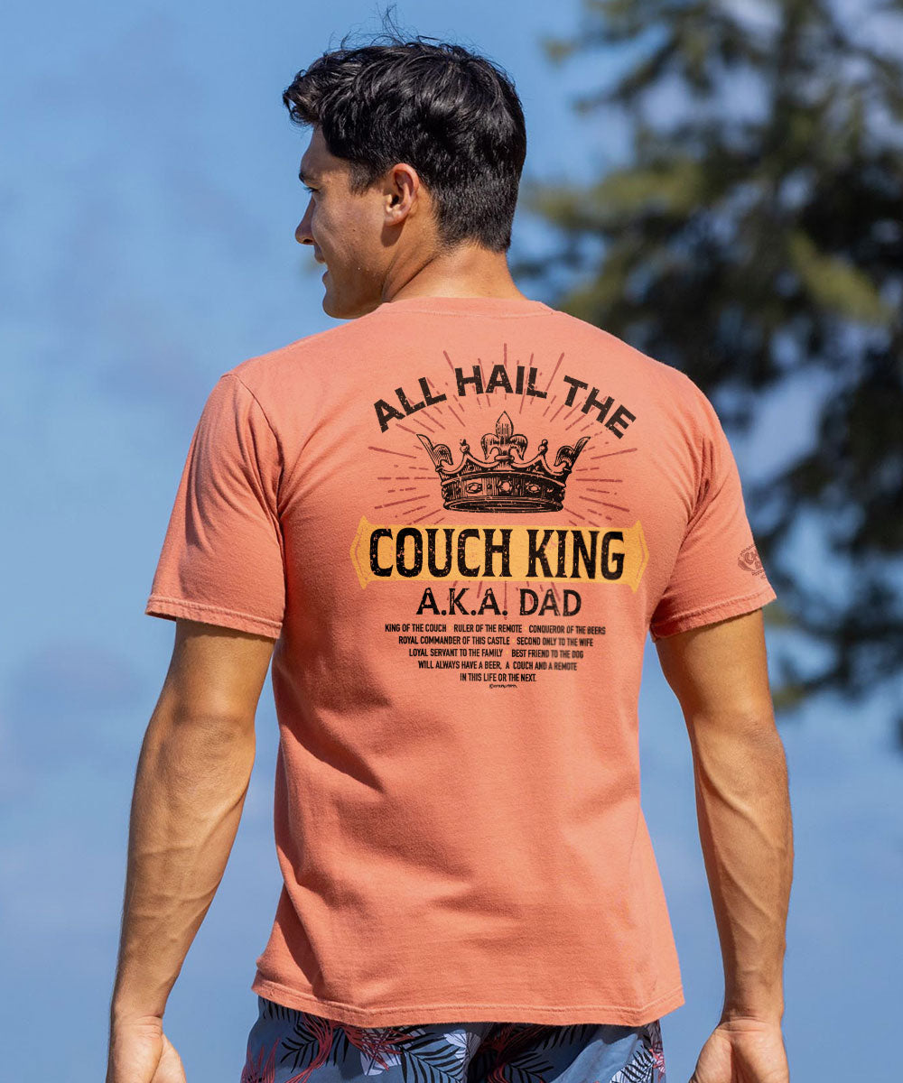 A man wearing a brown short sleeve crewneck t-shirt with the text 'ALL HAIL THE COUCH KING A.K.A. DAD' printed on the back. View 2