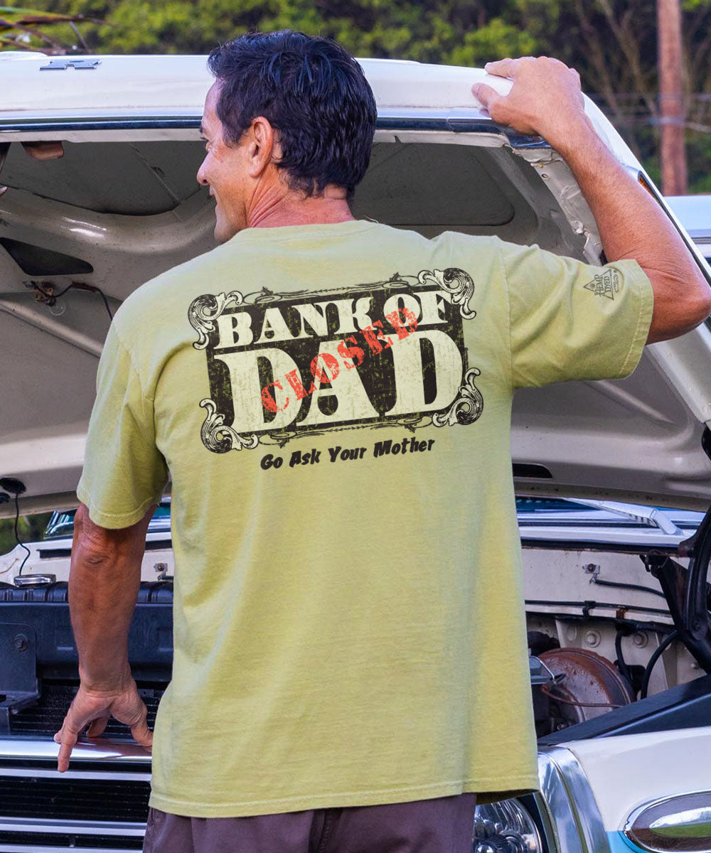 A person wearing a green short sleeve crewneck t-shirt with 'HANK OF DAD' printed on the back in bold letters, in front of a car. View 2
