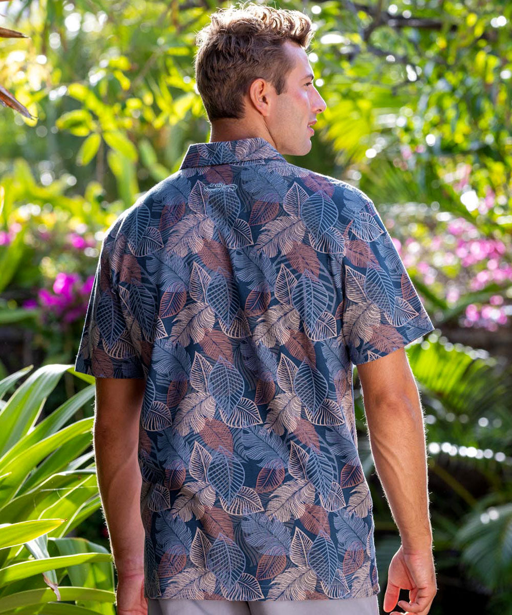 Man wearing a blue leaf-patterned shirt standing in a garden with greenery and flowers. View 3