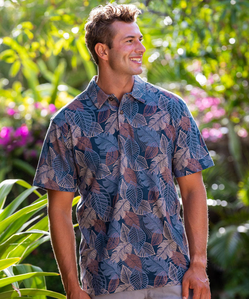 Man wearing a blue leaf-patterned shirt standing in a garden with greenery and flowers. View 2