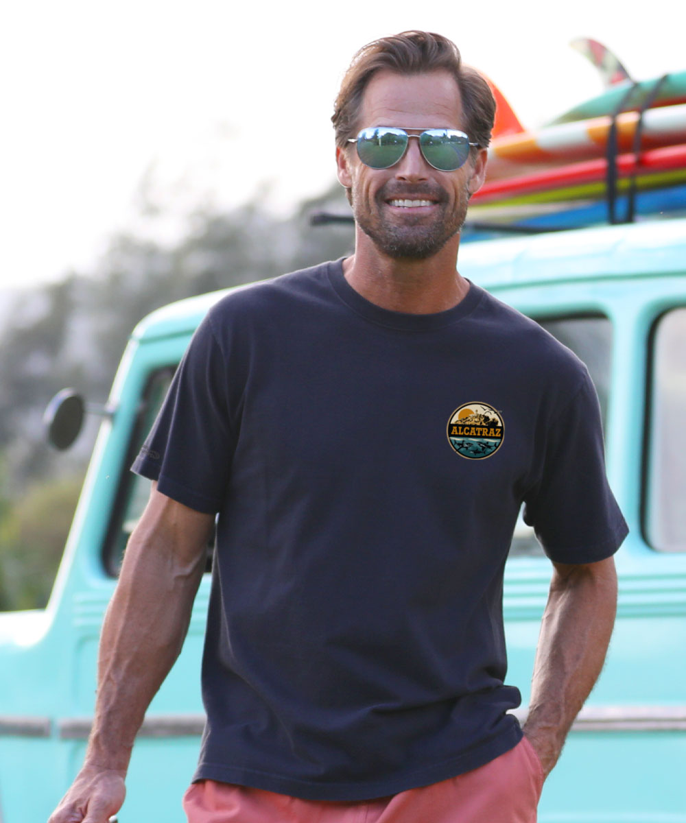 Man wearing a navy blue t-shirt with a logo, standing in front of a turquoise van with surfboards. View 3
